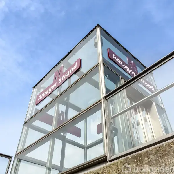 Glashus indeholdende en elevator i bevægelse, omgivet af lys og blå himmel. Skilte med teksten "Amager Strand" er monteret på bygningen.