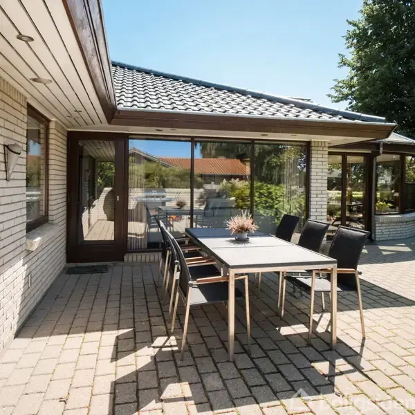 Terrassemøbler står stille på en brolagt gårdsplads ved et murstenshus med store vinduer. Omgivet af grønne buske og træer under en klar blå himmel.