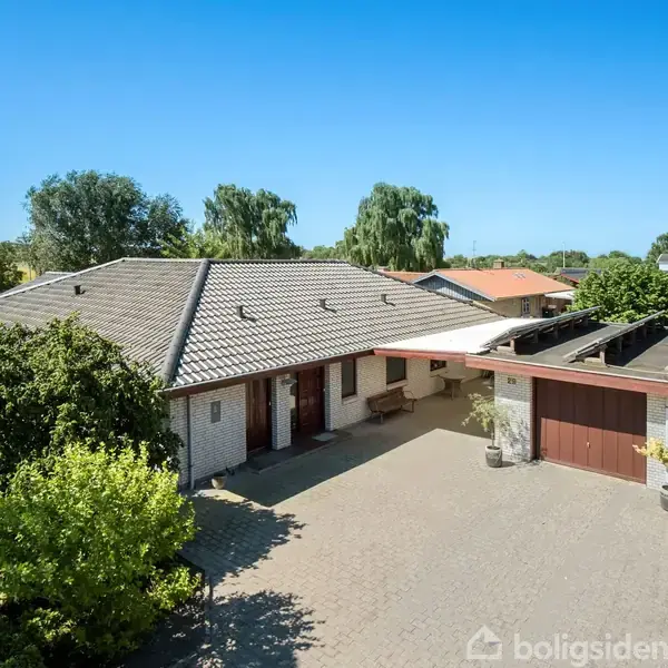 Huset står stille med grå tagsten, omgivet af en stor flisebelagt gårdsplads og træer i baggrunden. Til højre ses en overdækket garage.