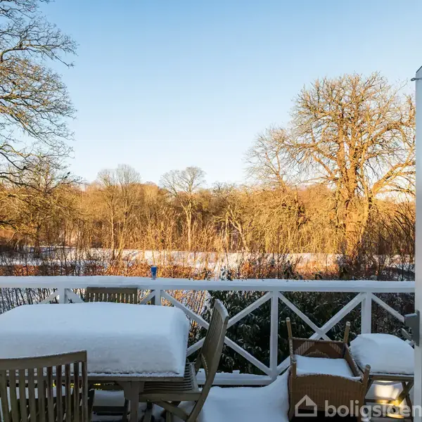 Et snedækket terrassebord står ubenyttet på en veranda. Bagved ses frostklædte træer og en snedækket mark oplyst af sollys.