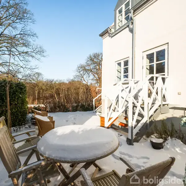Rundt havebord dækket af sne står med stole omkring i en sneklædt have. En trappe fører op til et hus med hvide vinduer. Løvfældende træer i baggrunden under blå himmel.