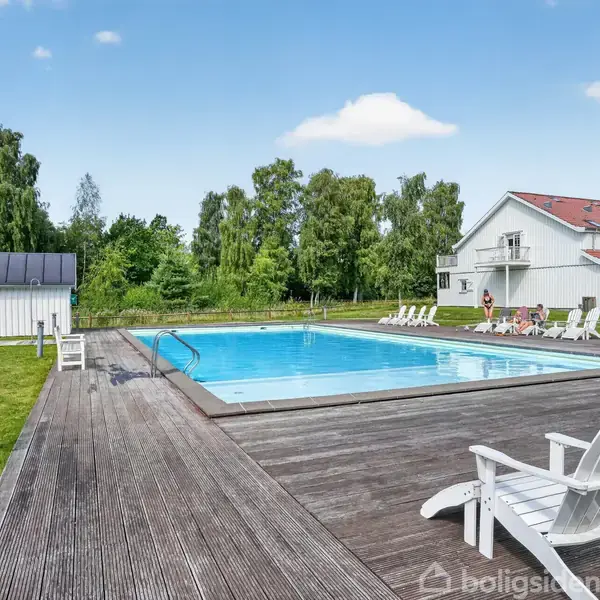 En stor udendørs swimmingpool med træterrasse omgivet af grønne træer. Flere hvide liggestole står omkring poolen, og et hvidt hus ses i baggrunden.