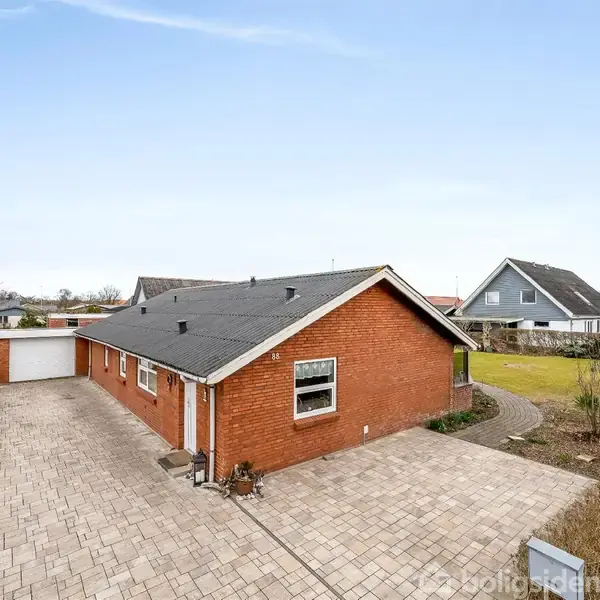 Rødt murstenshus står stille ovenpå en stor, flisebelagt indkørsel. Bag huset ses en garage, mens nabohuse og en velplejet have omgiver området under en blå himmel.