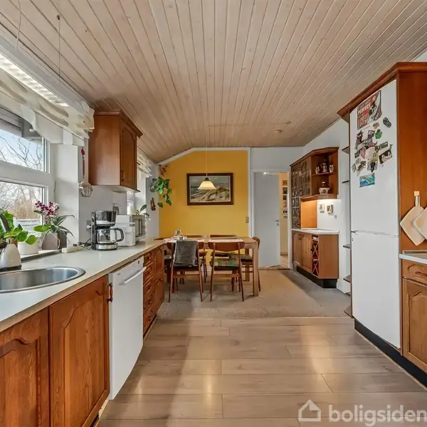 Køkken i træ med vask og komfur til venstre, køleskab til højre. Et spisebord med fire stole står foran en gul væg med et maleri. Vinduer til venstre.