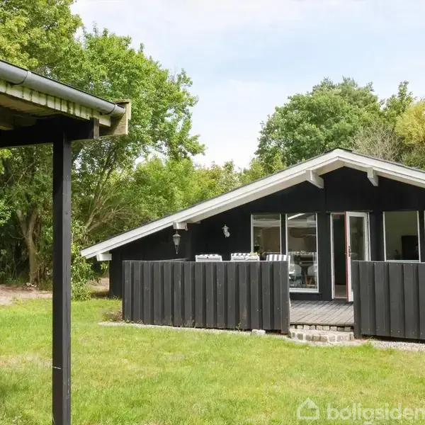 Et sort træhus med terrasse står blandt grønne træer og græsplæne. Store vinduer åbner til naturen, omkranset af en lav, sort stakit.