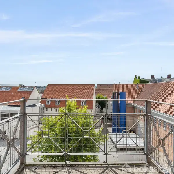 Altan med metalgelænder rækker ud fra en bygning med udsigt over hustage med røde tegltage. En grøn trætop er synlig under altanen. Klar blå himmel i baggrunden.