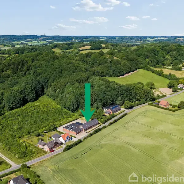En pil peger mod en rækkehusbygning, der ligger ved siden af åbne marker, omgivet af skovklædte bakker og spredte huse i et landligt område.