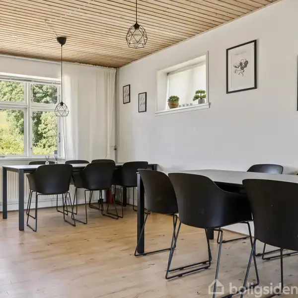 Spisebord med sorte stole står i et lyst rum med trægulve og træloft. Væggene har ophængte billeder og planter på en hylde. Store vinduer giver udsigt til grønt område.