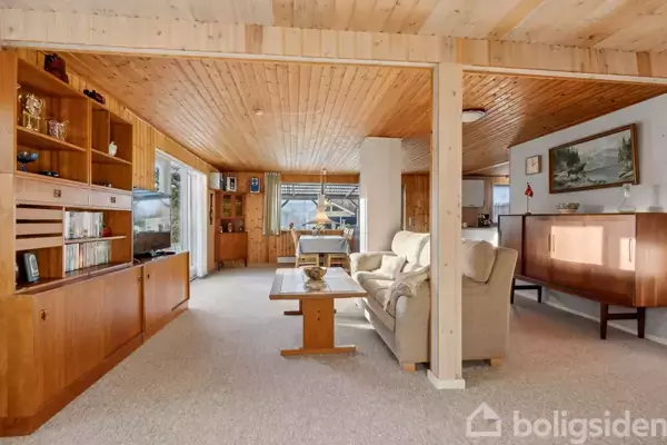 Stue med lyse trævægge og loft, brunt reolsystem langs venstre væg, beige sofa og sofabord i centrum; i baggrunden ses spisebord og flere vinduer.