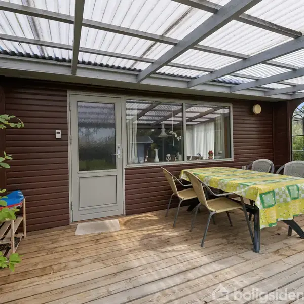 Overdækket terrasse med træbord dækket af ternet dug. Omgivet af stole, brune trævægge og ruder, skaber en hyggelig siddeplads udendørs. Oval vinduesdetalje og grønne planter tilføjer charme.