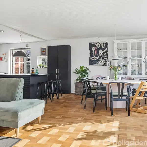 En stue med en grøn sofa og et træspisebord med sorte stole. I baggrunden er et moderne køkken med sorte skabe. Rummet har et lyst parketgulv og store vinduer.