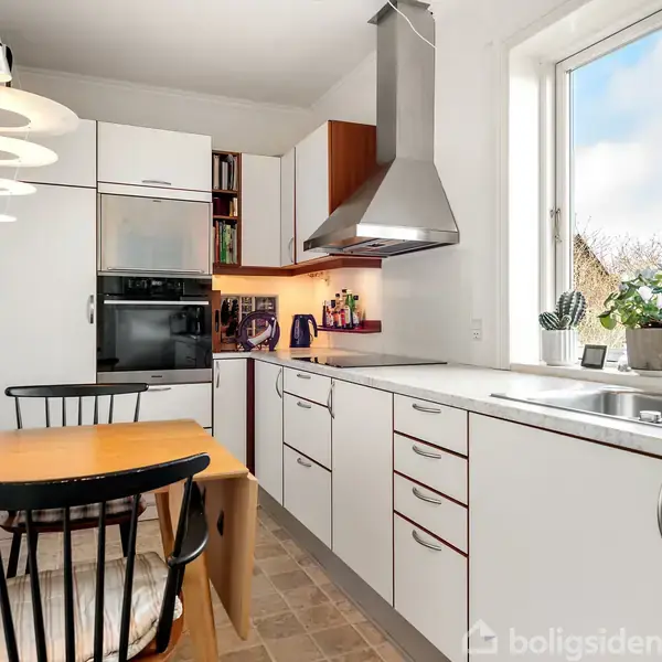 Køkken med hvidt inventar langs væggen, bord med stol i forgrunden. Vinduet giver udsigt til natur, mens lamper hænger over bordet. Skabe og ovn er modernistiske i design.