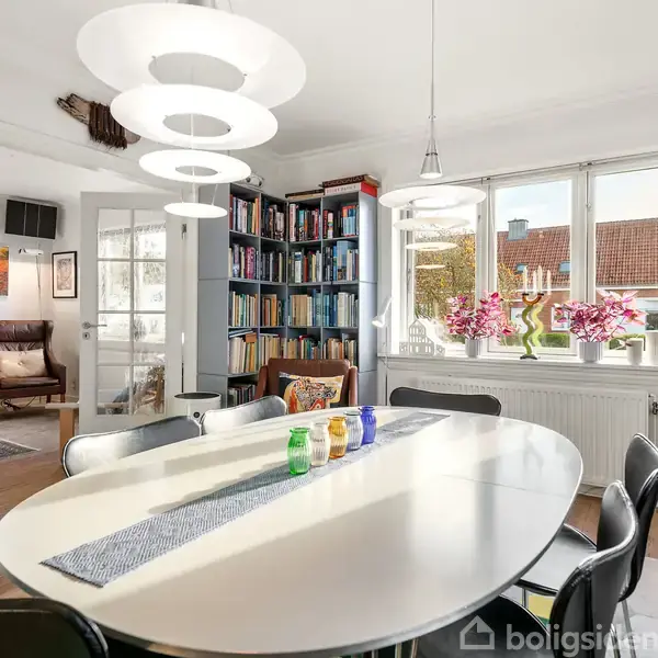 Ovalt spisebord med seks stole står i en lys spisestue. Bordet er pyntet med farvede glas og en løber. I baggrunden er der en bogreol og vinduer med udsigt.