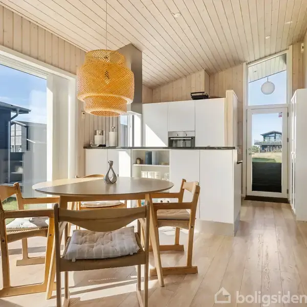 Et rundt spisebord med fire stole står i et lyst køkken med træloft og store vinduer. En flettet lampe hænger over bordet. Reolerne ved siden af er fyldt med bøger.