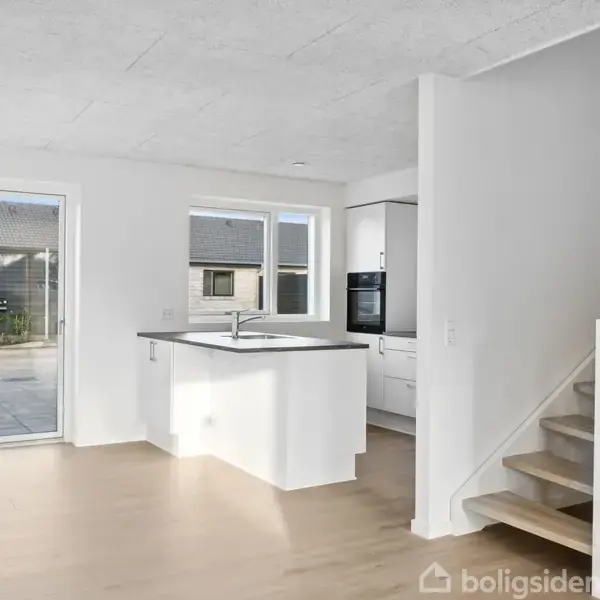 Et lyst køkken med en ø i centrum, placeret ved siden af en trappe. Et vindue og en dør åbner ud til en terrasse i baggrunden.