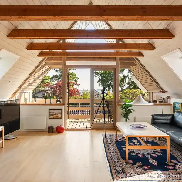 Stue med sort lædersofa og træbord på tæppe. Store træbjælker i loftet. Store vinduer med udsigt til grøn have og terrasse. Tegninger og lamper dekorerer rummet.