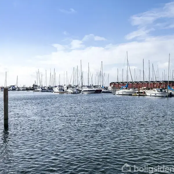 Både ligger fortøjet i en marina, omgivet af vandoverfladen og pæle. I baggrunden ses en række røde bygninger og en blå himmel med hvide skyer.
