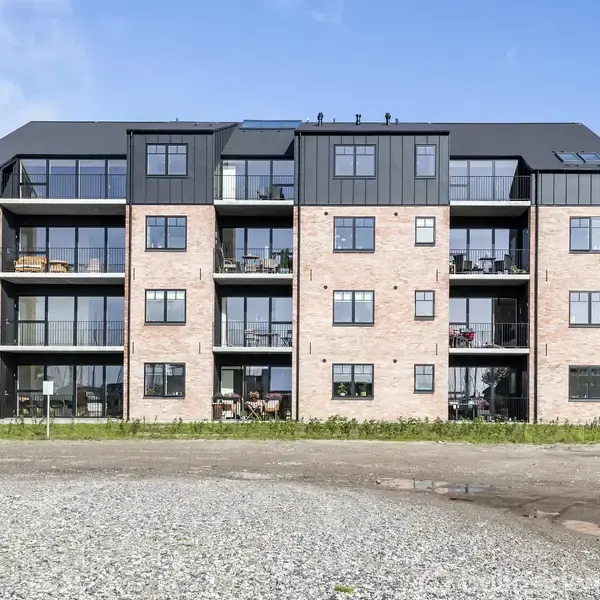En moderne fire-etagers lejlighedsbygning ses med store glasbalkoner og sort tag. Bygningen står på en grusplads og er omgivet af nogle andre bygninger og grønne træer i baggrunden.