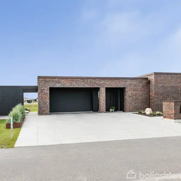 Moderne etplanshus i røde mursten har en stor indkørsel og tilstødende garage. Det ligger i et åbent, grønt boligområde med velplejede plæner og en blå himmel i baggrunden.