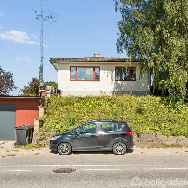 Et hvidt hus med røde vinduer står bag en garage og en sort bil parkeret på vejen. Der er en grøn skråning foran huset og en stor træ ved siden af.