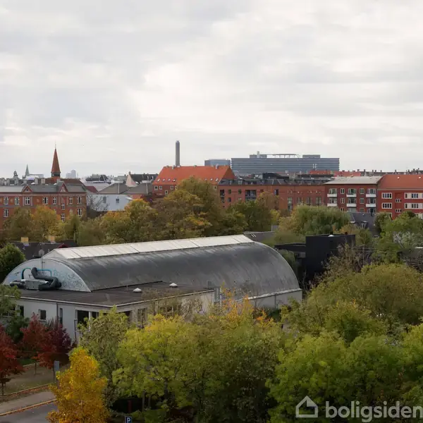 En hvælvet bygning omgivet af træer står stille i en by med røde hustage og kirketårn, under en overskyet himmel.