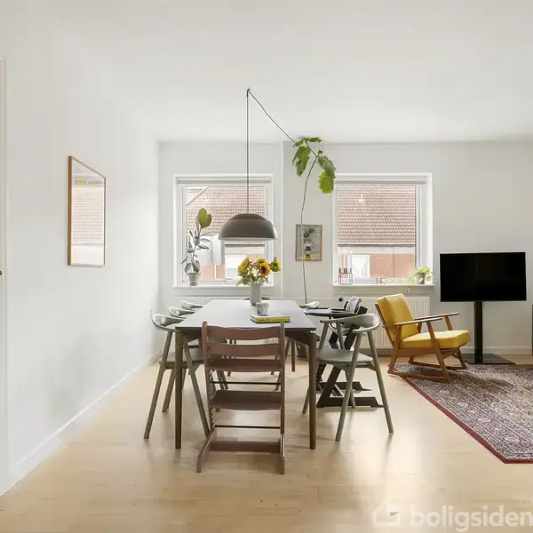 Spisebord med stole er dækket med tallerkener og en vase med blomster, placeret i en stue med sofa, lænestol og tæppe. Store vinduer giver naturligt lys.