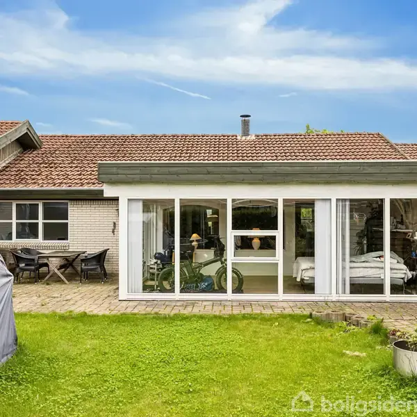 Et hus med en stor udestue har en glasfacade, der viser indre møbler og planter. Foran er en velplejet græsplæne og nogle blomsterkrukker i et roligt boligområde.