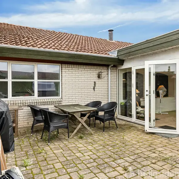 En terrasse med et bord og fire sorte stole står på en belagt plads. Til venstre er en grill, og i baggrunden ses et hus med en glasdør indtil.