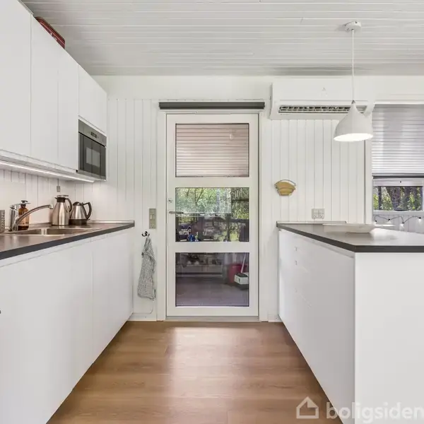 Køkken, minimalistisk stil, har hvide skabe, grå bordplader, indbyggede apparater og en grå tekande. Trægulv og loftslampe tilføjer varme. En glasdør og vindue med udsigt udenfor.