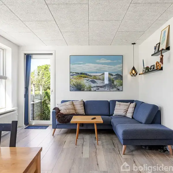 Blå sofa står i en stue med lyse vægge og trægulv. Et maleri hænger over sofaen. Rummet har store vinduer og en glasdør, der leder ud til en have.