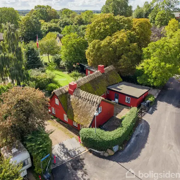 Rødt stråtækt hus omgivet af frodig have med høje træer. Beliggende langs stille vej i landsbyområde. Tydelige grønne områder og nærliggende huse.