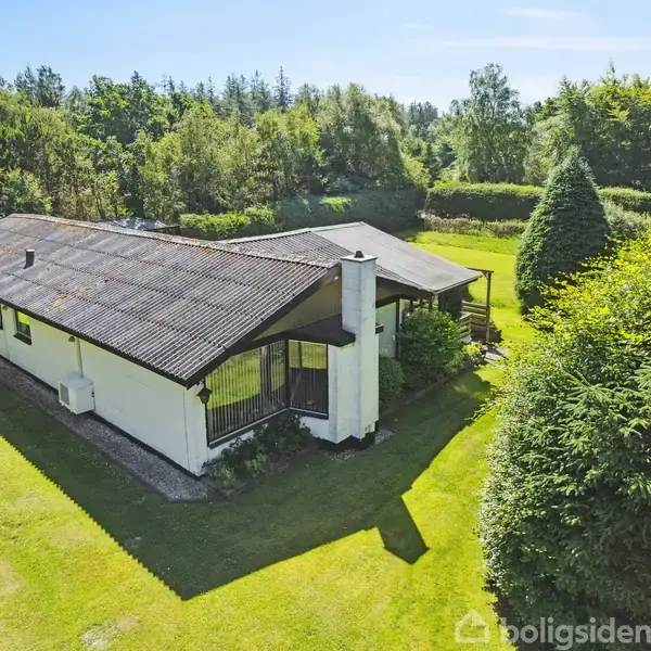 Et hvidt enfamiliehus med sort tag ligger omgivet af en velplejet græsplæne og tætte buske. Huset har en pejs og er placeret i en skovklædt, grøn omgivelser.