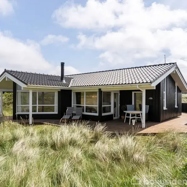 Sort hus med terracotta-tag står stille i et åbent landskab med højt græs og blå himmel. En enkelt terrasse med møbler ses foran huset.