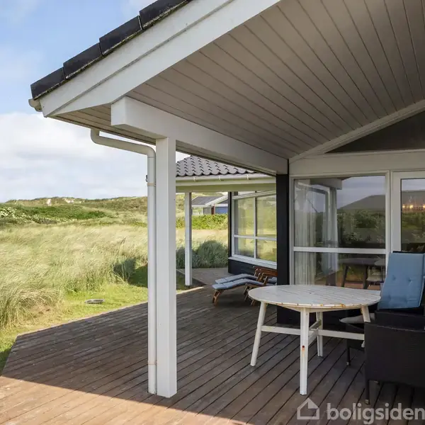 Terrassen har et rundt bord og en liggestol. Trædækket strækker sig ud under et halvtag. Omgivet af grønne bakker og græs i en åben, naturlig landskabsscene.