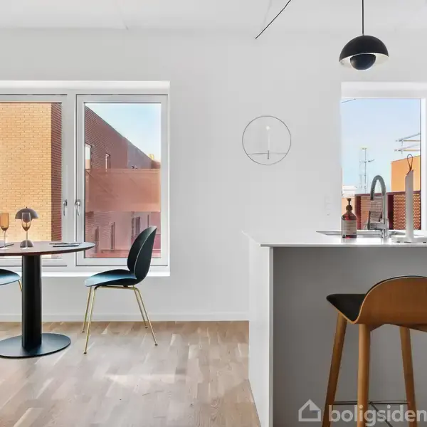 Et moderne spiseområde har et rundt bord med to stole foran store vinduer, der giver udsigt til rødbrune murstensbygninger. En høj barstol står ved en køkkenø med enkel indretning.