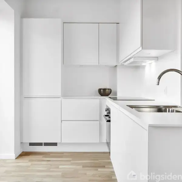 Hvidt køkken med indbyggede skabe og skuffer, en vask ved en grå bordplade og flaske. Trægulv og åbent vinduesparti til venstre, lys og moderne atmosfære.