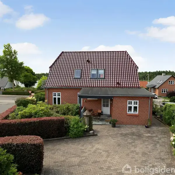 Rødt murstenshus med skråt tegltag har en forhave med hæk og indkørsel af fliser. Det står i et beboelseskvarter med lignende huse og grønne områder.