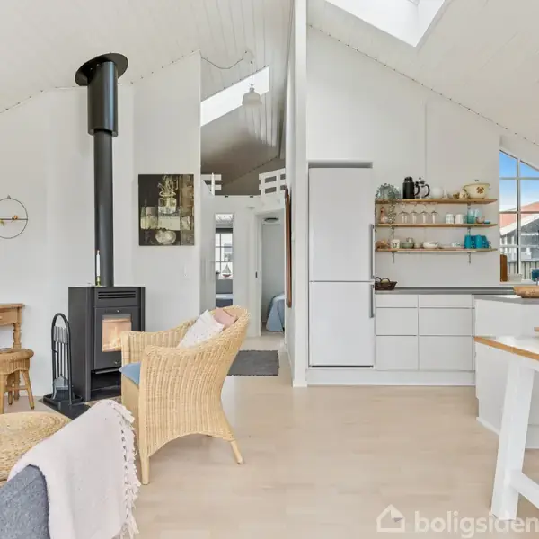 Stue med pejs, hvor kurvestol er midtpunktet. Ved siden af er et køkken med åbne hylder og komfur. Store vinduer giver lys, og et tv hænger på væggen.