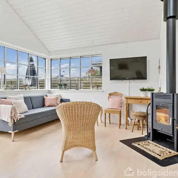 En moderne stue har en brændeovn tændt i forgrunden. En grå sofa med lyserøde puder står i hjørnet ved vinduerne. Et fjernsyn hænger på væggen.