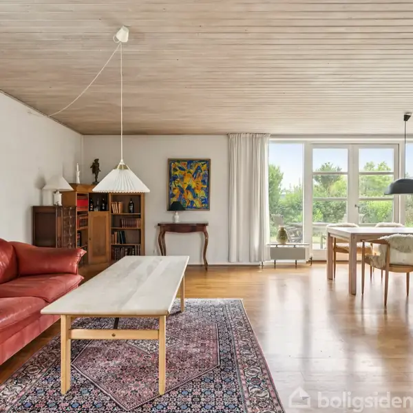 Rød sofa står ved træbord i stue. Der er kunst på væggene, bogreol og store vinduer med udsigt til have. Tæppe under bord, tv i højre side.