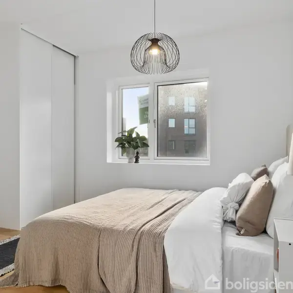 Soveværelse med en dobbeltseng dækket af en brun sengetæppe, ved siden af et hvidt natbord og en lampe. Vindue med plante, lyst hængelampe loftlys, skydedørsskab og trægulv.