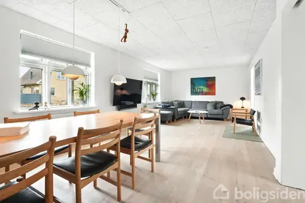 Spisebord med træstole står i moderne, lyst stueområde. Til venstre er vinduer og planter, mens en grå hjørnesofa med sofabord er til højre under et farverigt maleri.