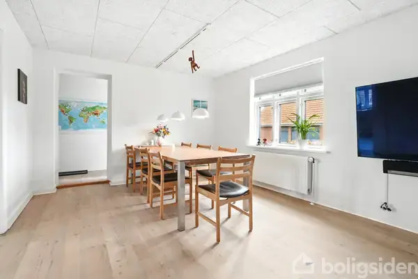 Et langt træbord med syv stole står i et lyst rum. En lampe hænger over bordet, og der er et vindue med en plante til højre. En verdenskortplakat ses i baggrunden.