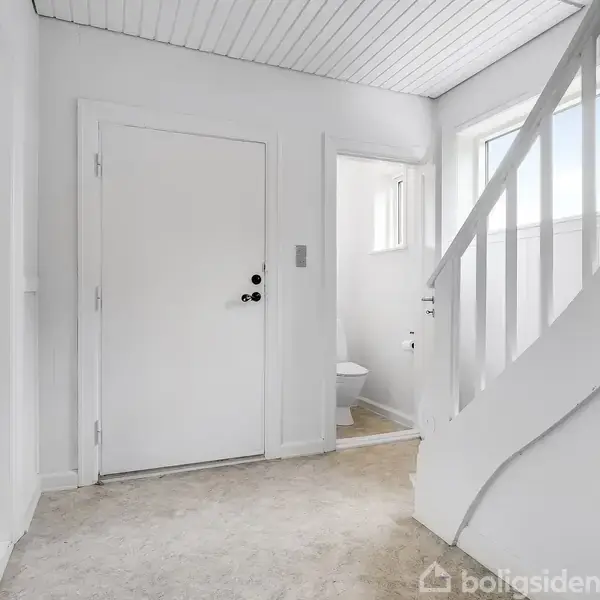 Dør står i en hvidmalet gang, med åbning mod et toilet. Til højre fører en hvid trappe til næste etage. Rummet har et lyst, minimalistisk udtryk.