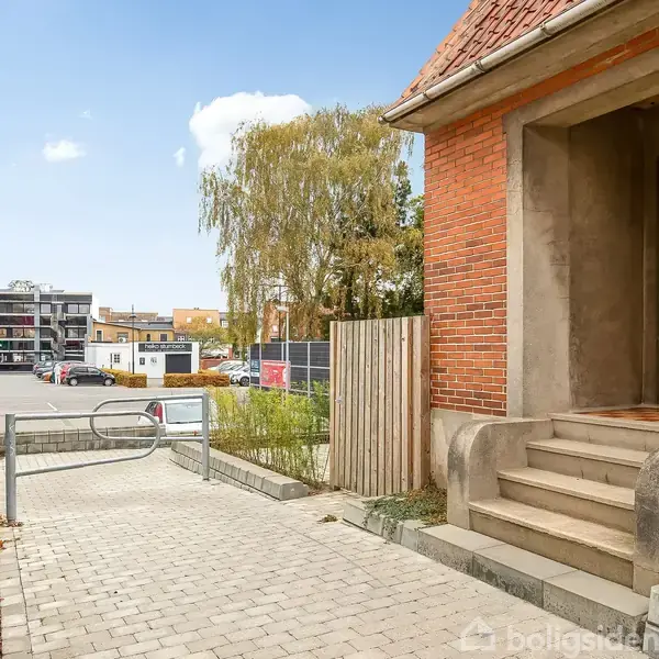 Rødt murstenshus med trappeopgang står ved siden af en brolagt sti. Baggrunden viser en urban gade med parkeringspladser og bygninger under en blå himmel.