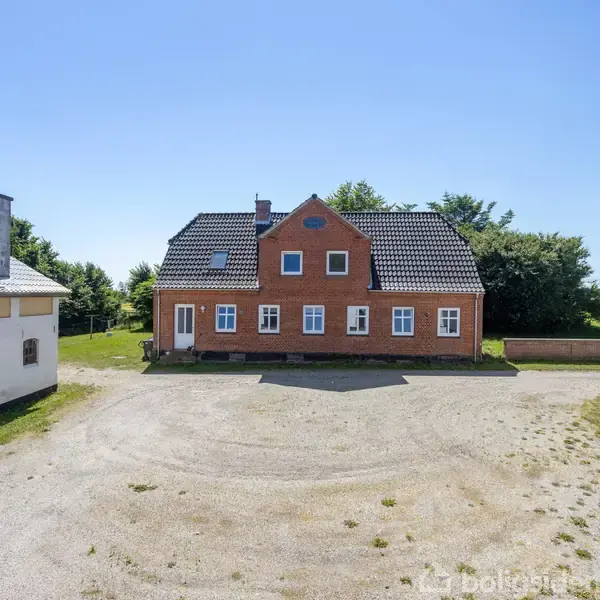 Rød murstenshus står centralt omgivet af to hvide staldbygninger med sorte tage. Husets vinduer vender mod en grusbelagt forplads med grønne marker i baggrunden under en klar blå himmel.
