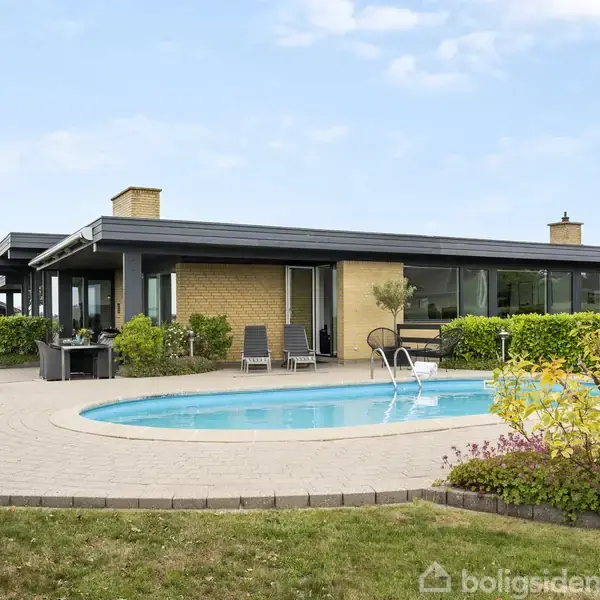 En moderne bolig med fladt tag omkranser en blå swimmingpool. Solstole og planter er placeret omkring poolen, mens haven er grøn og velplejet under en klar, blå himmel.