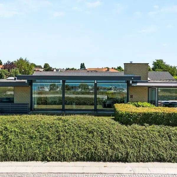 En moderne en-etagers villa med store glasfacader står bag en tæt, veltrimmet hæk. Huset er omkranset af en velholdt have med grønne buske og træer i et villakvarter.