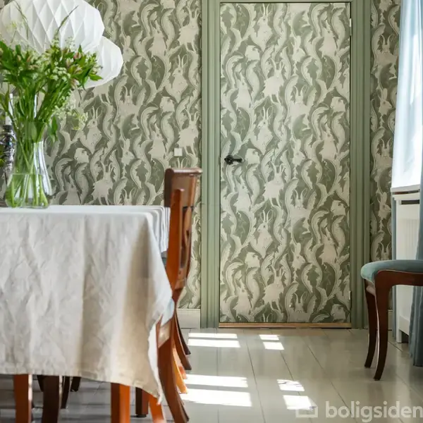En spisebord dækket med en hvid dug står i et rum med grønt-mønstret tapet. En vase med blomster er på bordet, og sollys skinner ind fra højre.