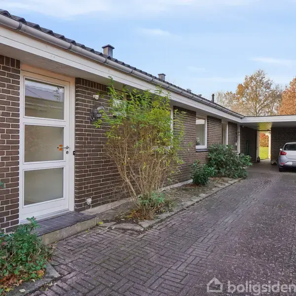 Et hus med mørk murstensfacade og en hvid dør står på højre side. En bil er parkeret under en carport til højre. Omgivet af buske og et flisebelagt indkørsel.